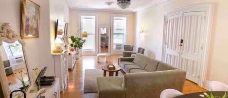 A spacious living room with a large beige sofa, a round coffee table, and a dining area with a small table and chairs. There is a large white door and a chandelier on the ceiling.