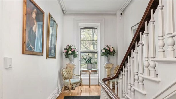 A hallway with a staircase, two chairs, a small table, and two flower arrangements.