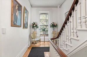 A hallway with a staircase, two chairs, a small table, and two flower arrangements. - Luxury Downtown Troy Apartments by Franklin Plaza. (Troy)