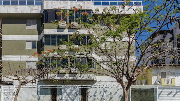 A multi-story residential building with balconies, a tree in the foreground, and a clear sky.