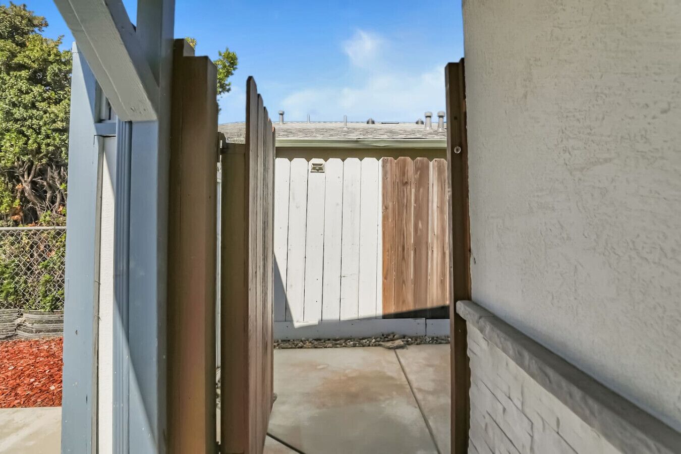 A narrow concrete pathway leading to a wooden gate, with a white wall on one side and a fence on the other.