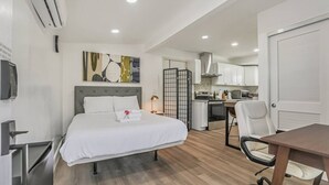 A modern hotel room with a bed, a desk, a chair, a kitchenette, and a TV.