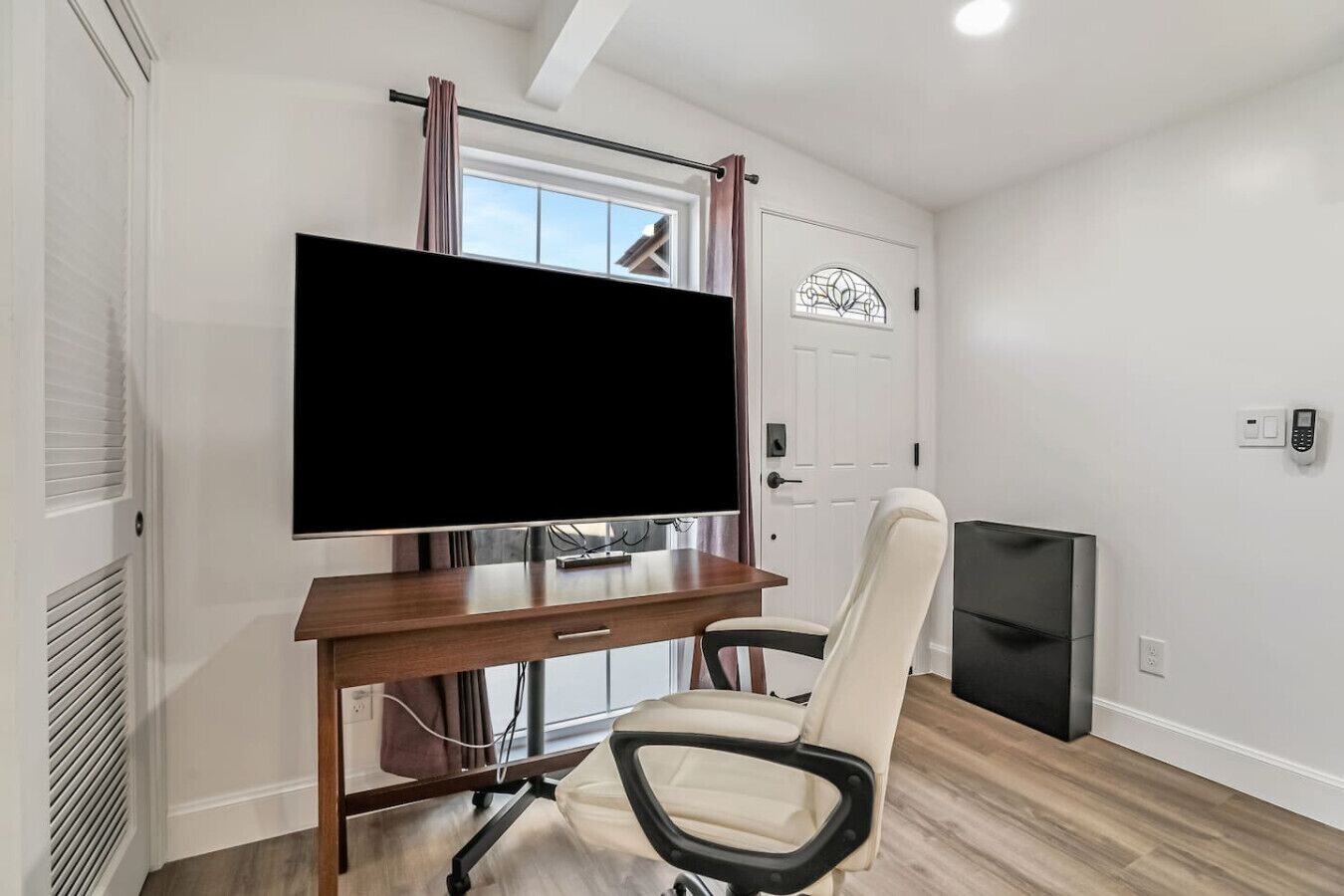 A room with a flat-screen TV mounted on a wooden stand, a white office chair, a door with a window, and a thermostat on the wall.