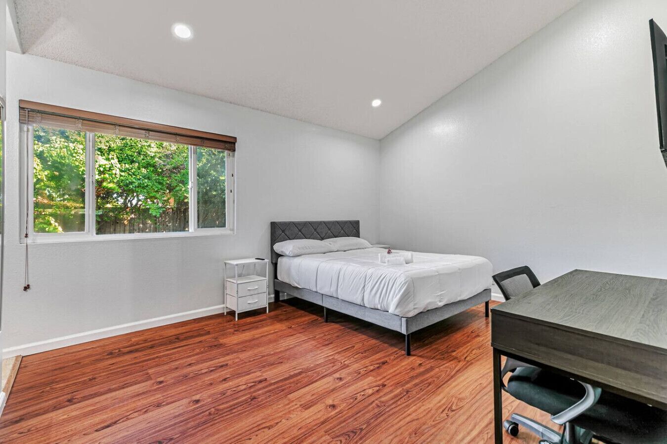 A bedroom with a bed, a desk, and a window with a view of greenery.