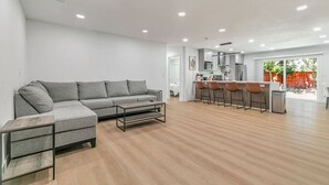 Una sala de estar moderna con un sofá seccional gris, una mesa de centro de madera y una cocina con barra alta y taburetes.