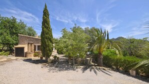 Un bâtiment rustique en pierre, doté d’une allée en gravier, entouré d’une végétation luxuriante et de grands arbres.