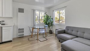 A compact living space with a kitchenette, a dining area with a glass table and chairs, a grey sofa, and a large window with a view of trees. - Downtown 1bd/1ba ADU (Sunnyvale)