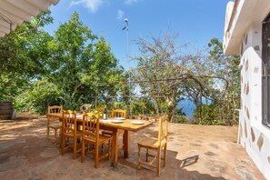 Zona d'exterior per dinar amb taula i caires, envoltada de vegetació i vista al mar.