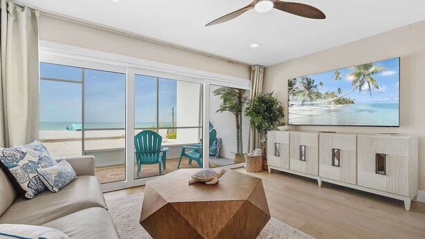 A spacious living room with a large TV, a wooden coffee table, and a view of the beach through large sliding doors. - Beachfront Paradise! Ground level with Heated pool (Holmes Beach)