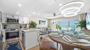 A modern kitchen with white cabinetry, a built-in microwave, and an oven. There is a dining area with a wooden table set for two, and a view of the ocean through large windows. - Beachfront Paradise! Ground level with Heated pool (Holmes Beach)