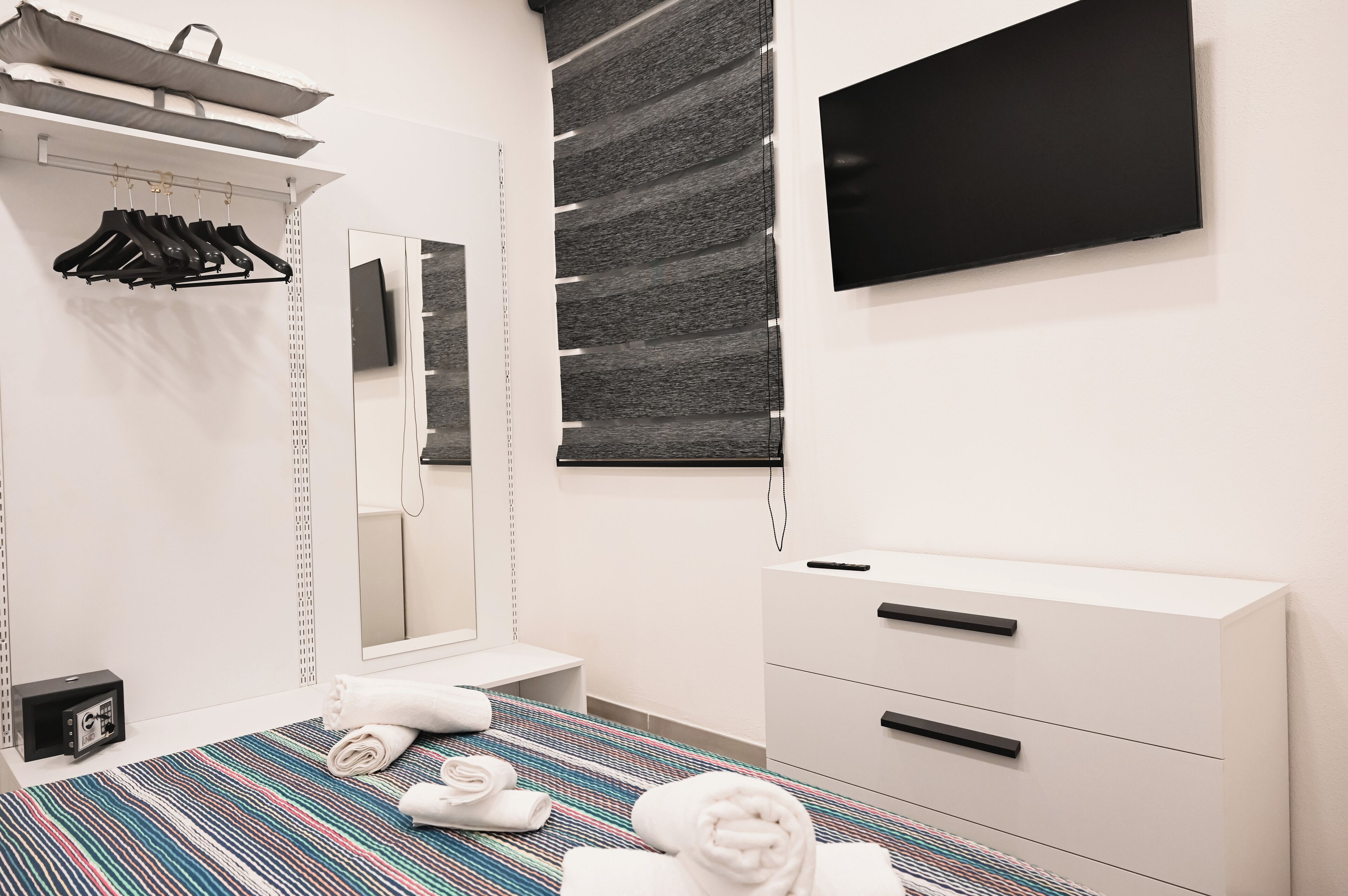 A modern hotel room with a bed, a bedside table, a wardrobe, a mirror, and a flat-screen TV mounted on the wall.