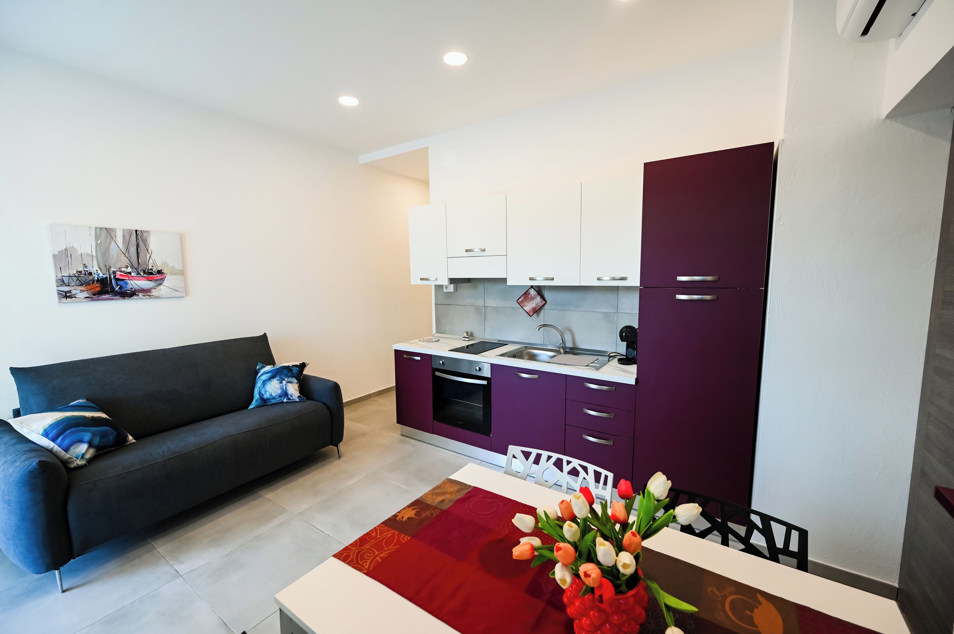 A modern living room with a sofa and a dining table with tulips. A kitchen with purple cabinets and white countertops is visible.