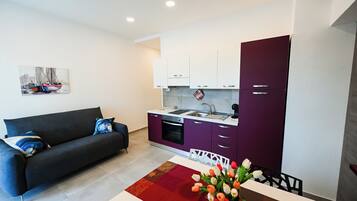 A modern living room with a sofa and a dining table with tulips. A kitchen with purple cabinets and white countertops is visible.