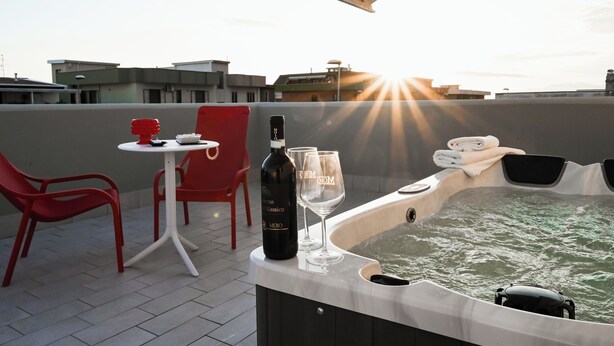 Une terrasse sur le toit avec un jacuzzi, des chaises rouges, une table, une bouteille de vin et des verres.