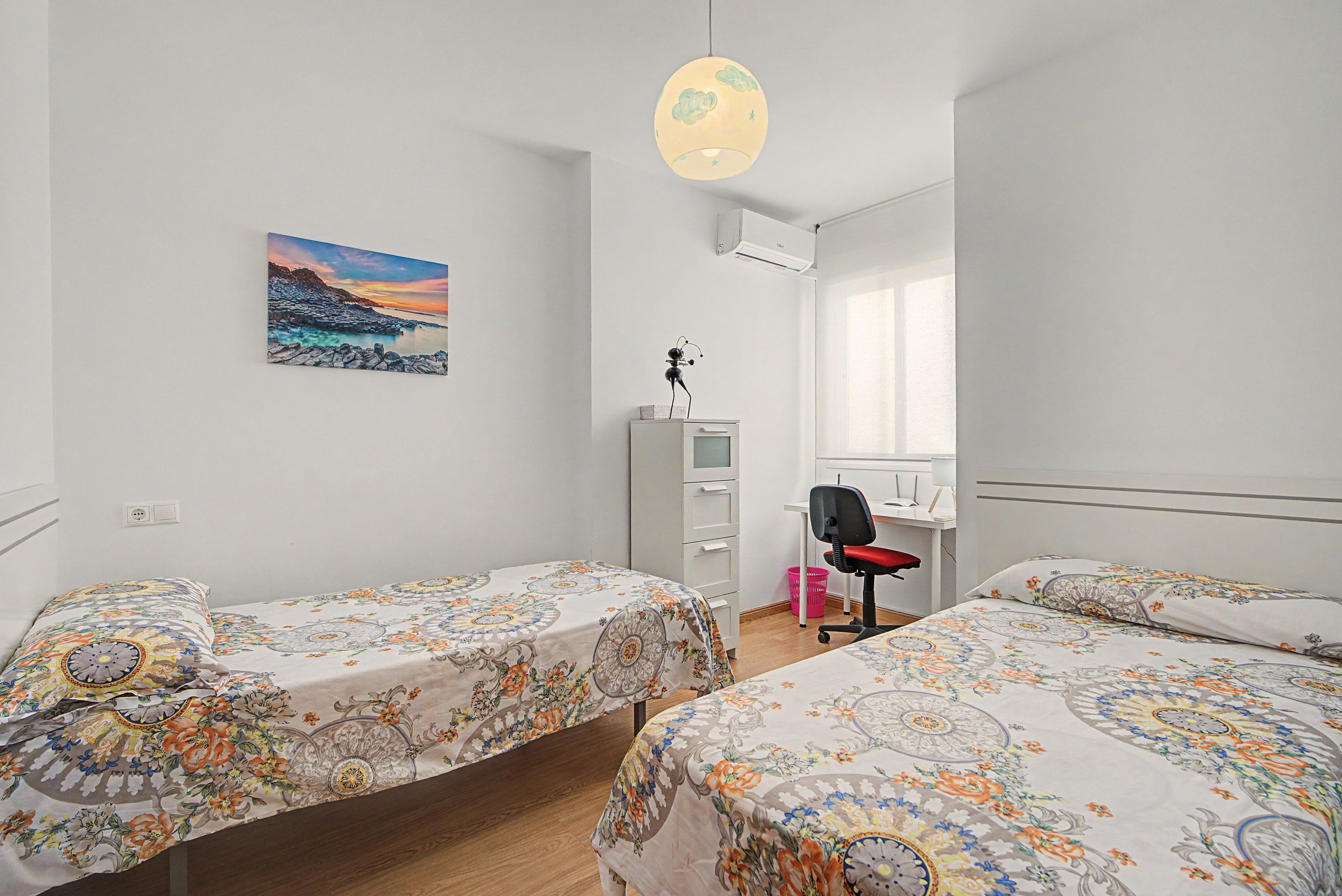 A bedroom with two beds, a desk, a chair, a wardrobe, and a painting on the wall.