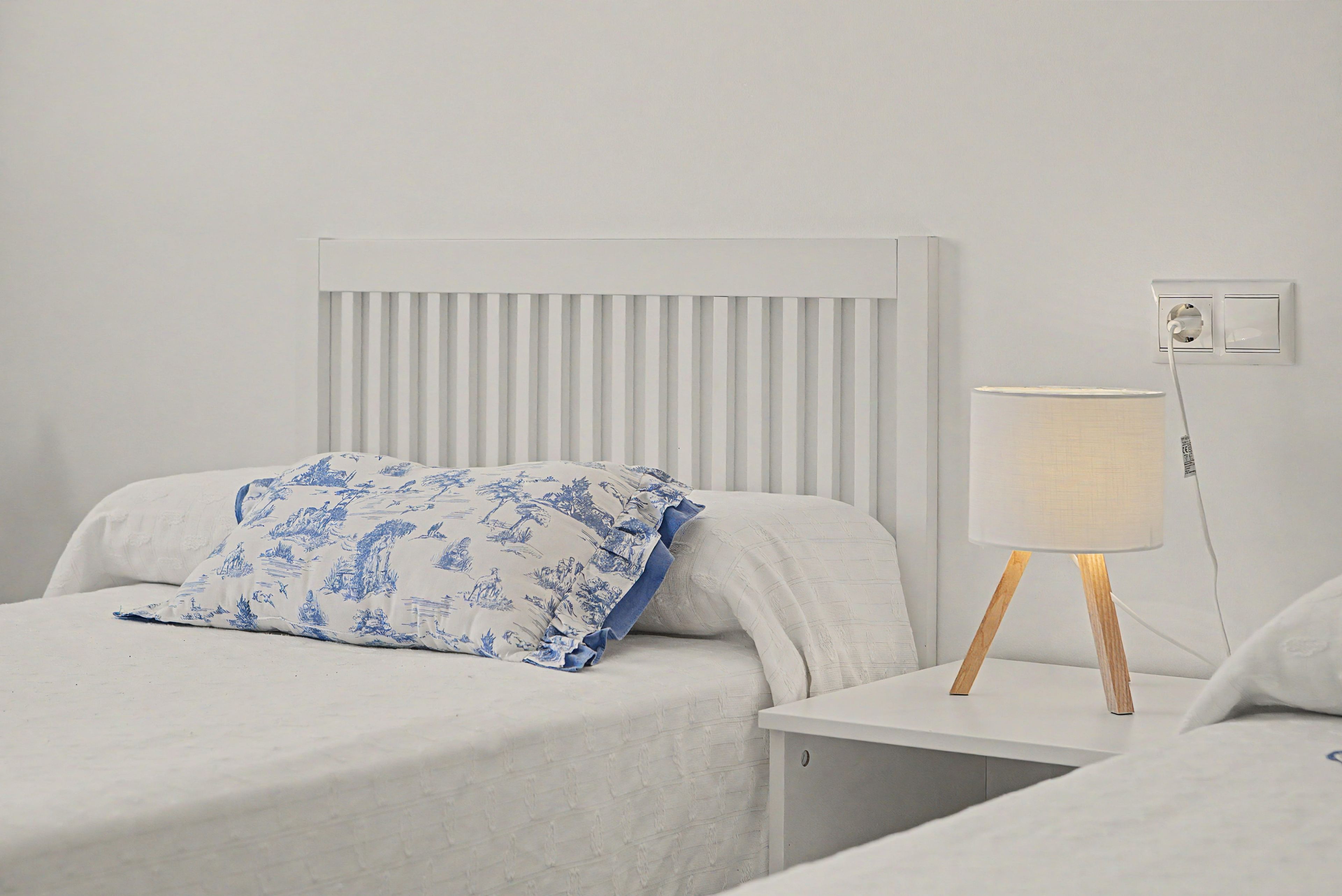 Una cama con cabecera blanca y almohada de estampado azul, una mesita de noche con lámpara y un enchufe en la pared.
