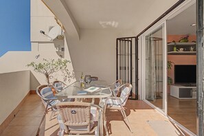 A glass-top table with wicker chairs, a wine bottle, and a glass on a balcony with a view of the sky and a building.