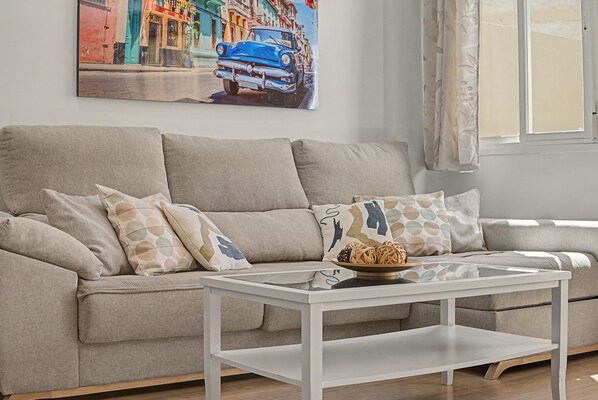 Una sala de estar con un sofá color beige, almohadones estampados, una mesa de centro blanca y un cuadro de un coche azul en la pared.