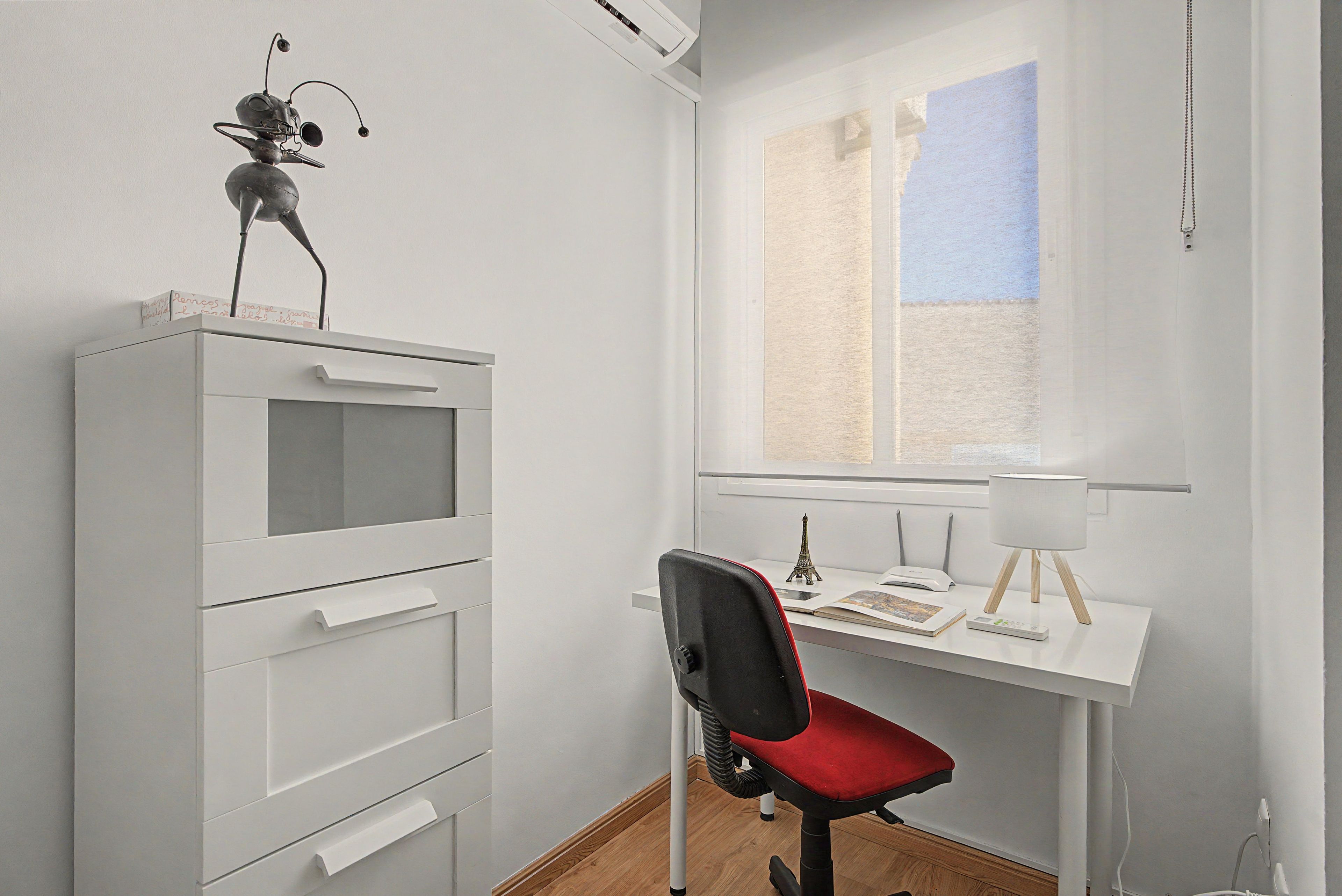 Una habitación con un escritorio blanco, una silla, un armario pequeño, una ventana y una lámpara.