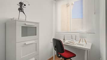 Una habitación con un escritorio blanco, una silla, un armario pequeño, una ventana y una lámpara.