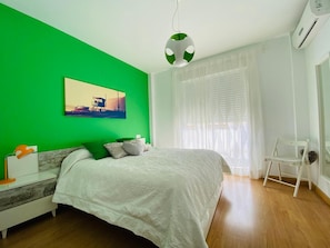A bedroom with a bed, a nightstand, a chair, a window with curtains, and a painting on the wall.