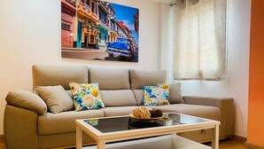 Un salon avec un canapé beige, deux coussins à motifs floraux, une table basse en verre et un tableau représentant un paysage urbain accroché au mur.