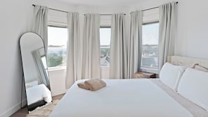 Une chambre d’hôtel moderne dotée d’un grand lit, d’un miroir en pied et offrant une vue sur la ville grâce à de grandes fenêtres.
