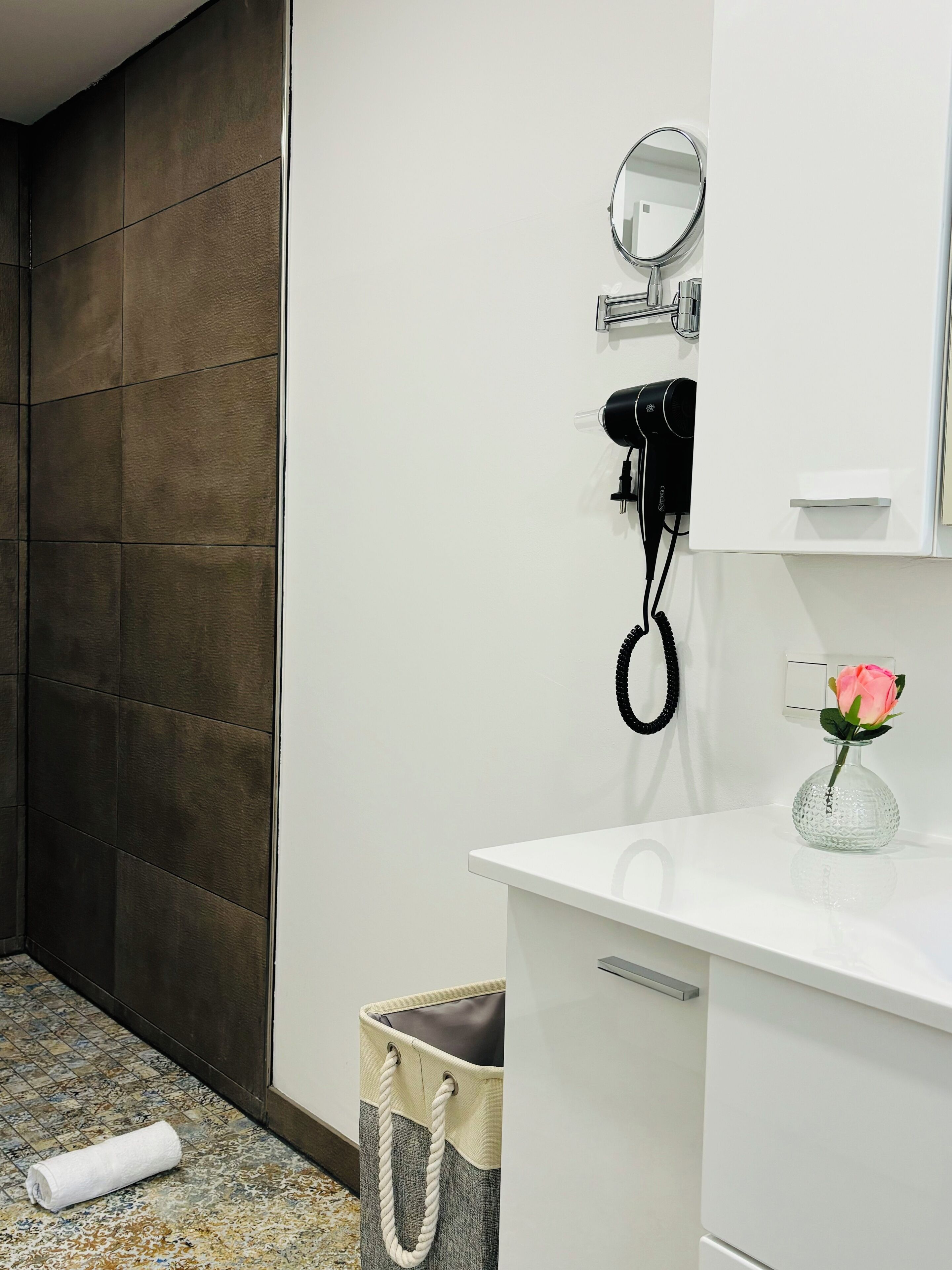 A bathroom with a white vanity, a round mirror, a hairdryer, and a vase with a pink rose.