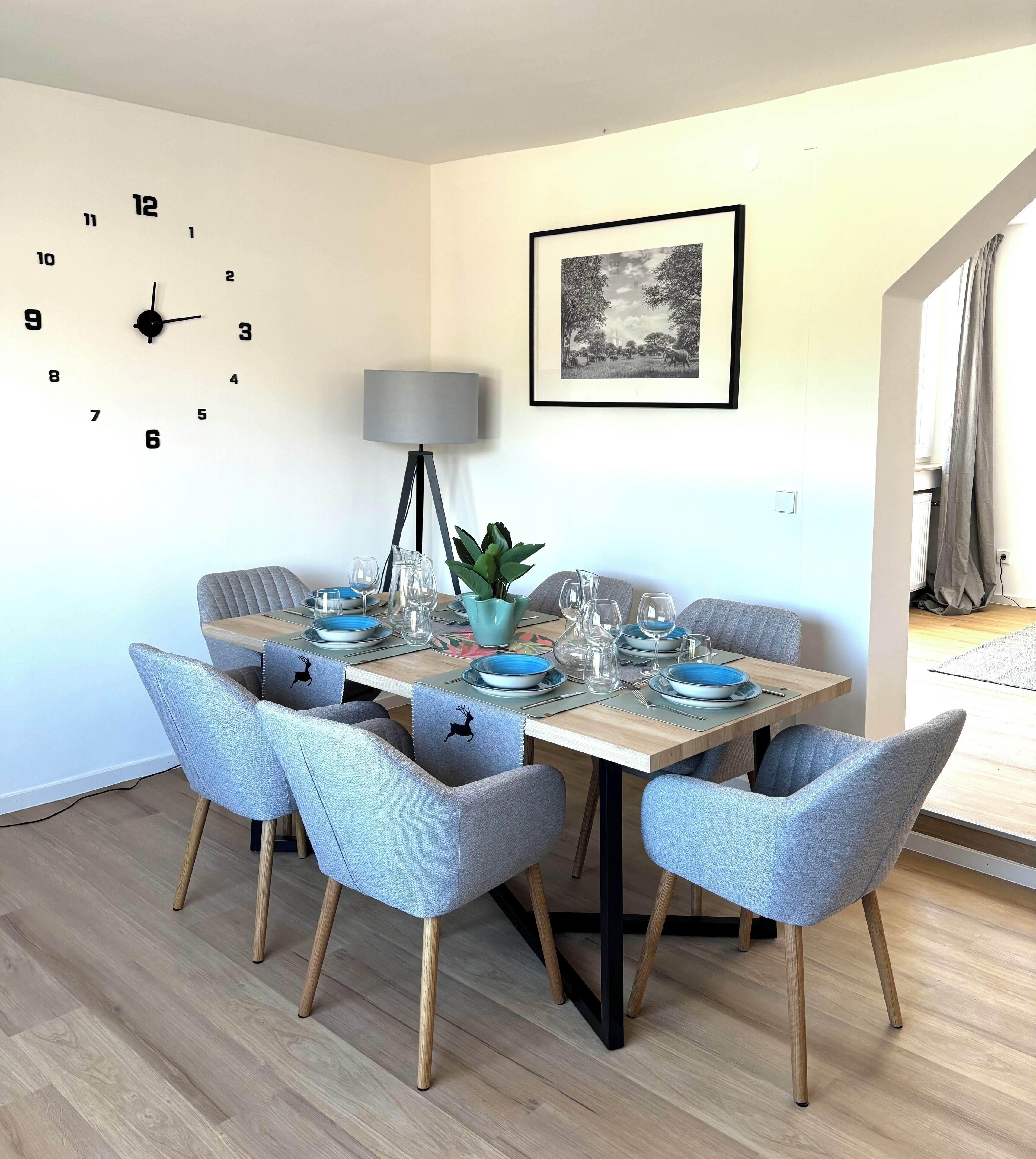 Ein Essbereich mit einem Holztisch, gedeckten Plätzen für vier Personen, grauen Polsterstühlen, einer Wanduhr, einer Stehlampe und einem gerahmten Bild an der Wand.