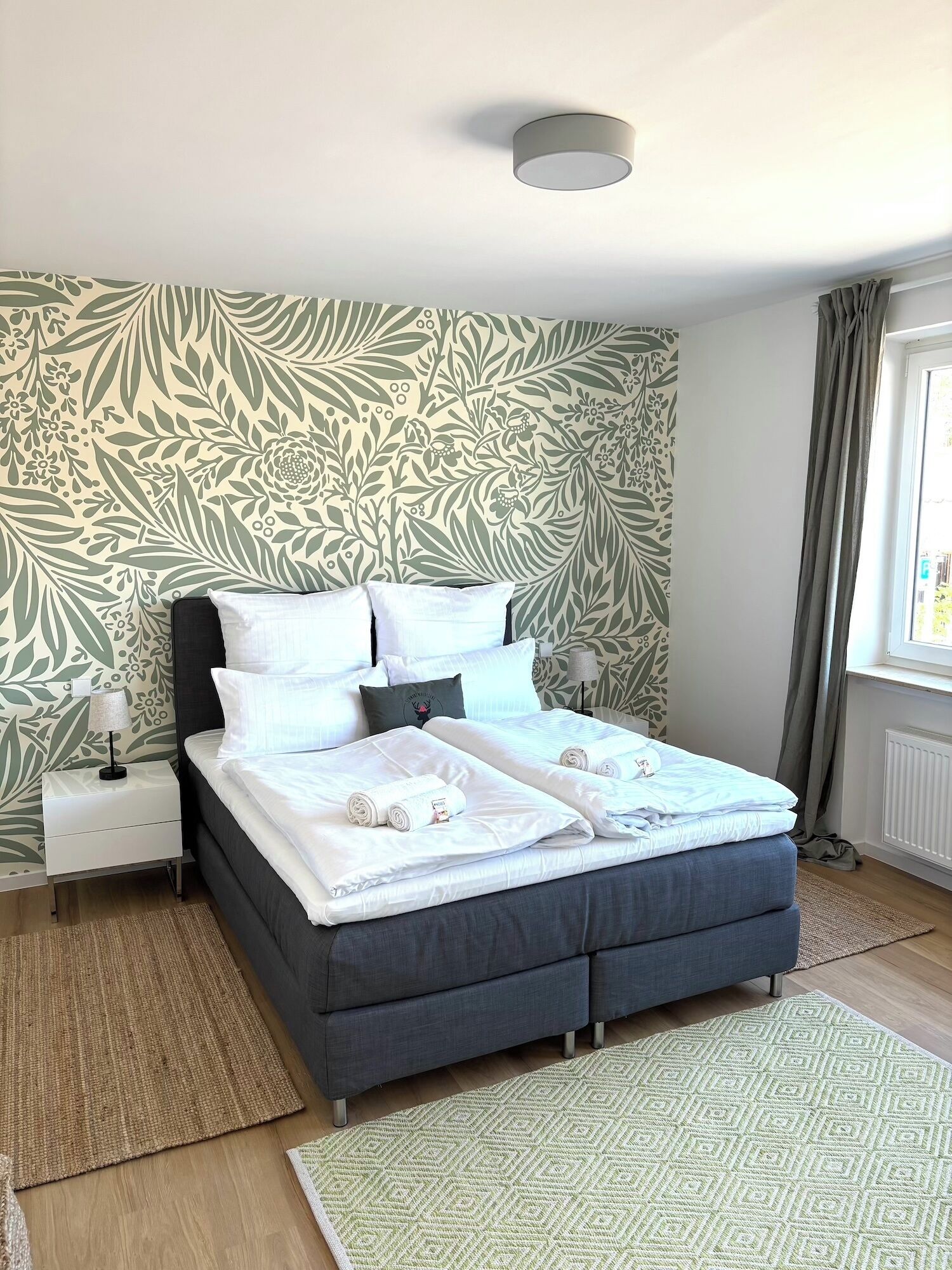 A double bed with a dark headboard, white bedding, and a patterned wall behind it. There is a small bedside table with a lamp, a window with curtains, and a patterned rug on the floor.