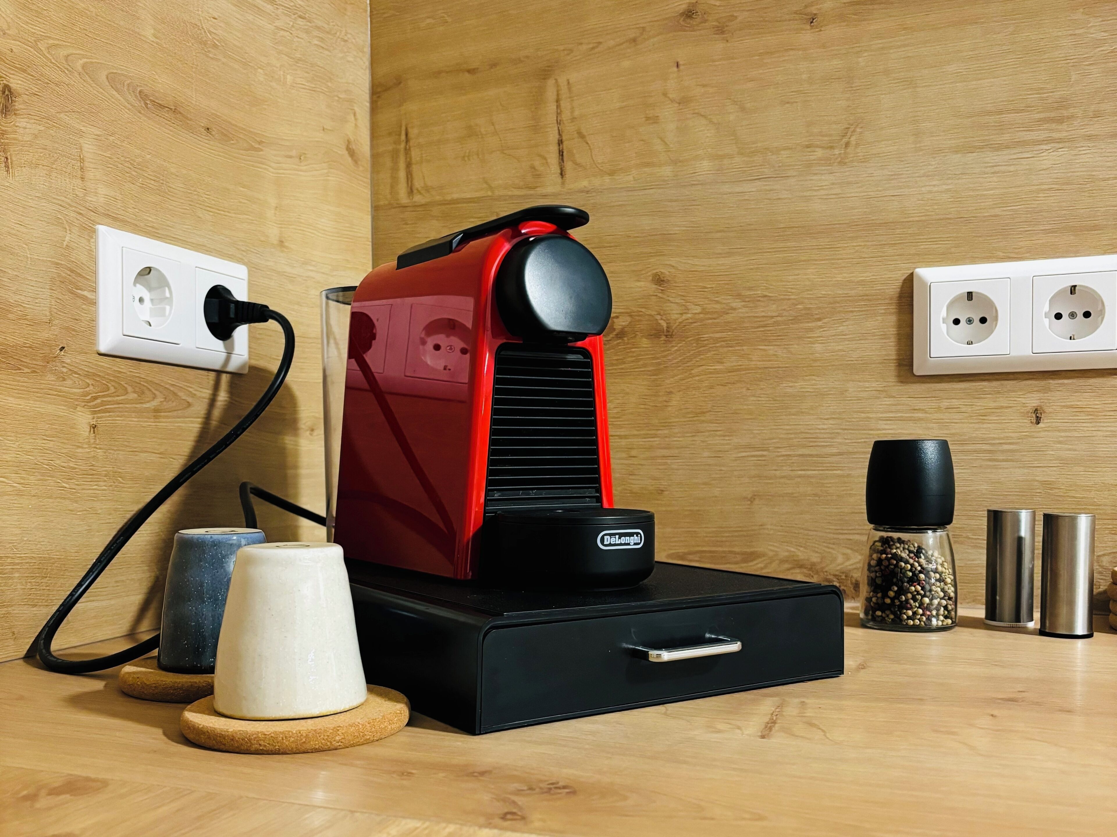 Una máquina de café sobre una mesa de madera, con un enchufe, un cajón y algunos recipientes de especias.