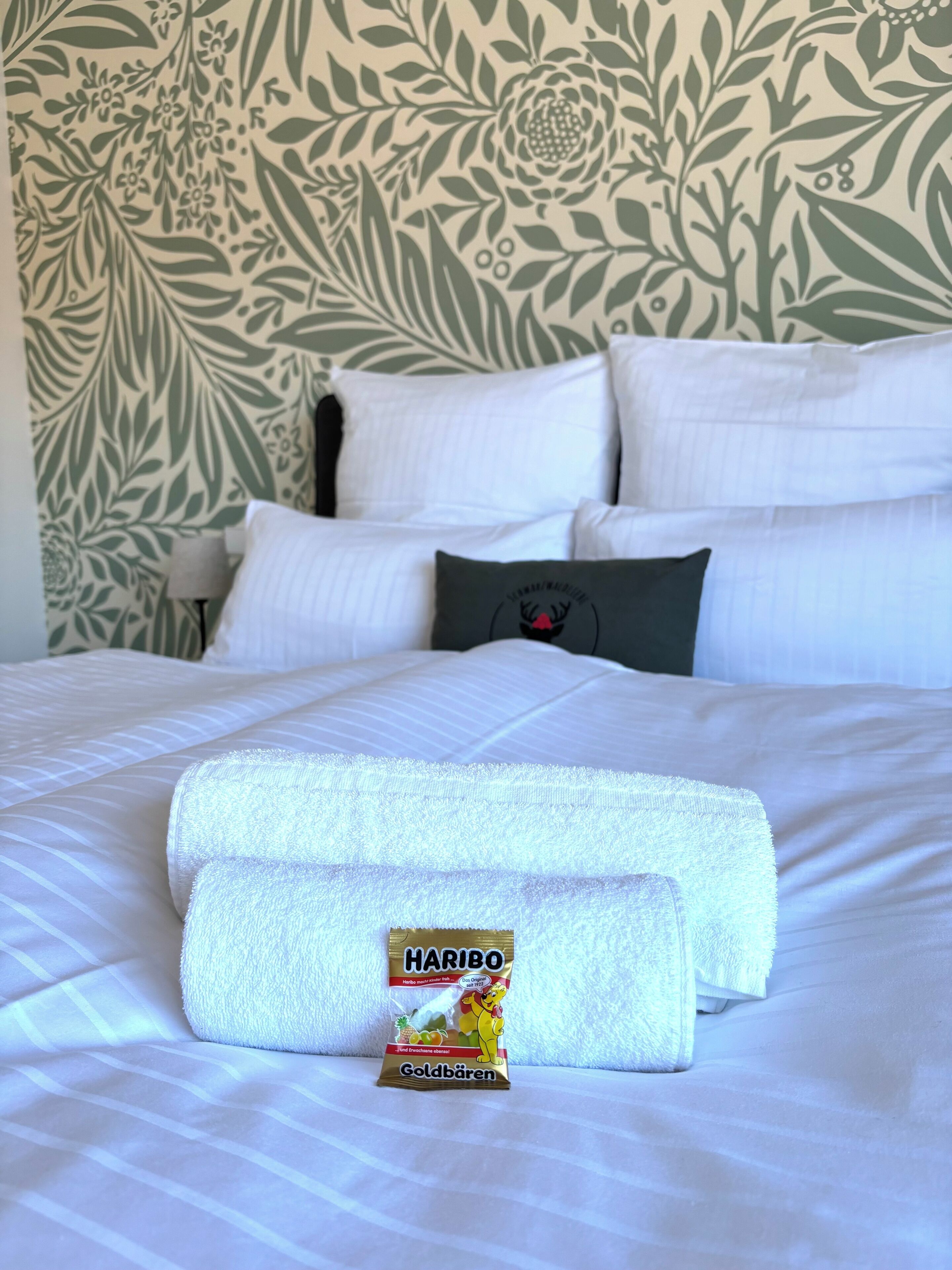 A neatly made bed with white linens and a folded towel, a Haribo Goldbären chocolate bar, and a pillow with a logo.
