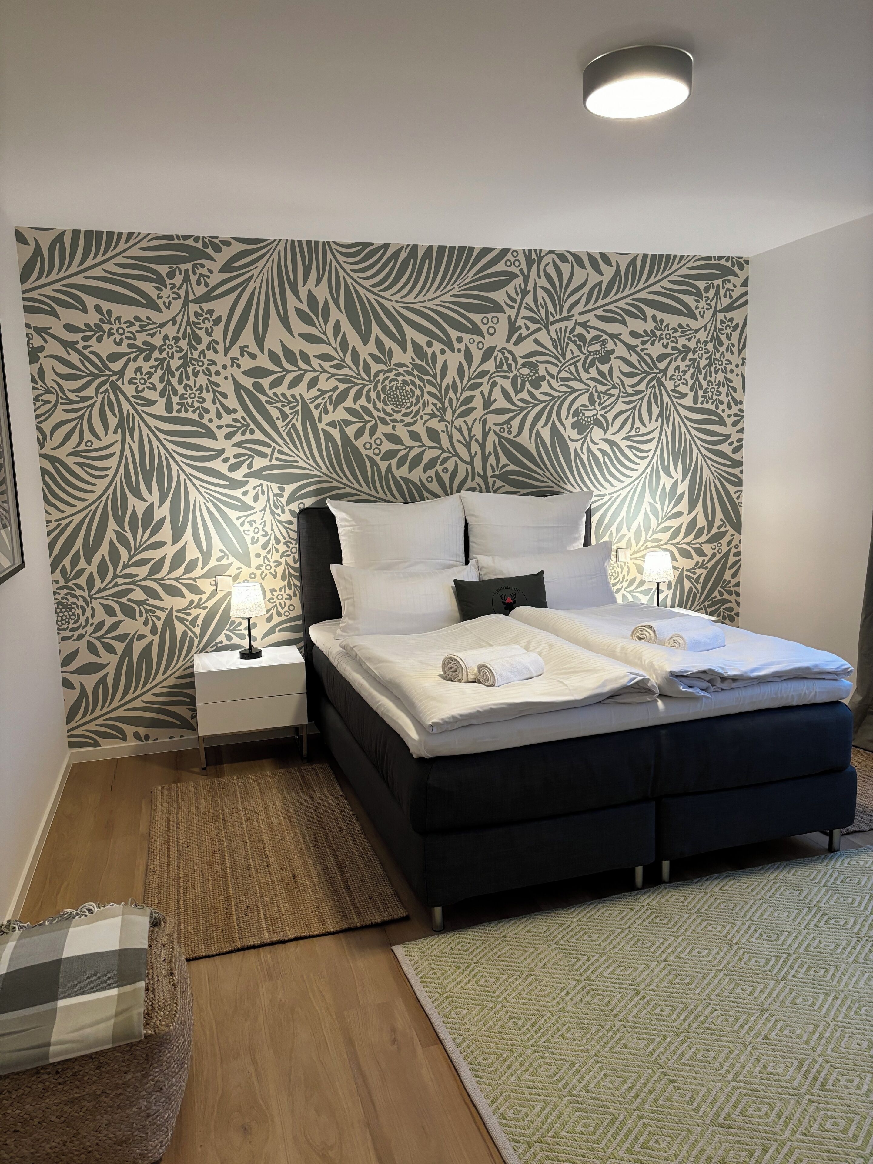 Een slaapkamer met een bed, nachtkastjes, een lamp, behang met een patroon en een geweven vloerkleed.