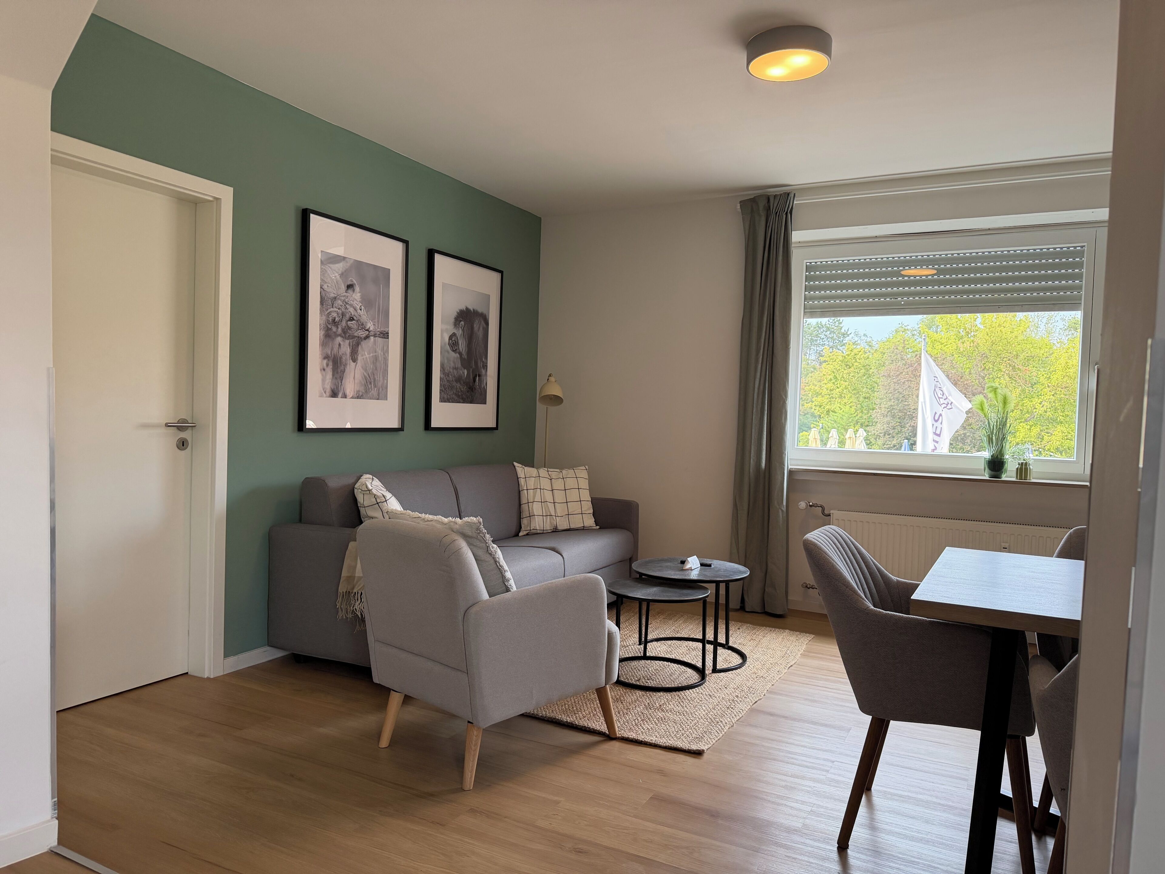 Una sala de estar con sofá, sillón, una mesa de centro y una mesa de comedor con sillas. Hay dos cuadros en la pared, una ventana con cortinas y una puerta.