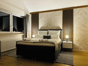 A neatly arranged hotel room with a large bed, a bedside table with a lamp, a window with curtains, and a wall with a floral pattern. - Rema Homes Bad Bellingen (Bad Bellingen)