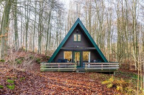 A cabin with a triangular roof, wooden deck, and a forest backdrop.