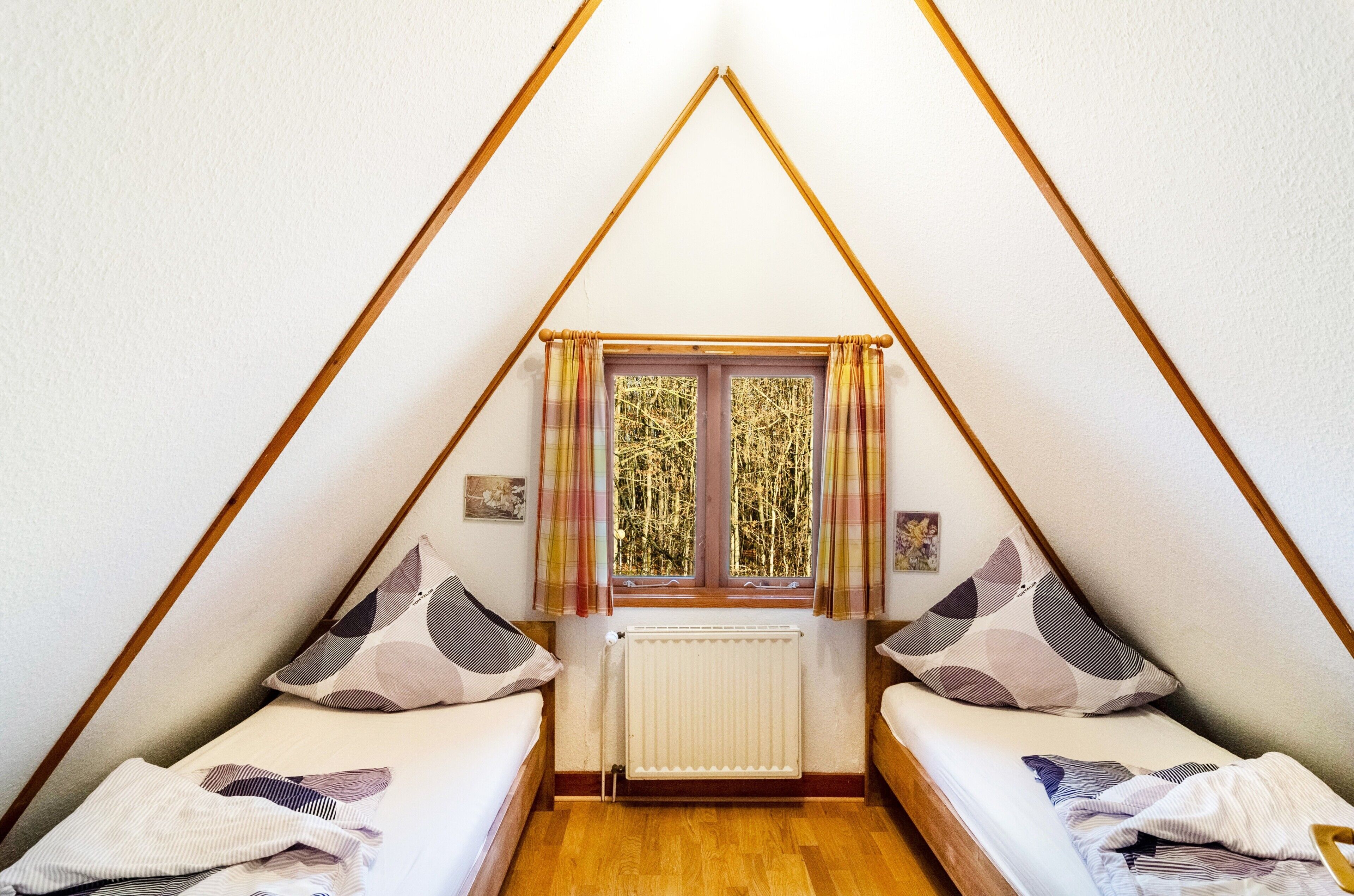Une pièce triangulaire avec deux lits, une fenêtre avec des rideaux, un radiateur et des poutres en bois.