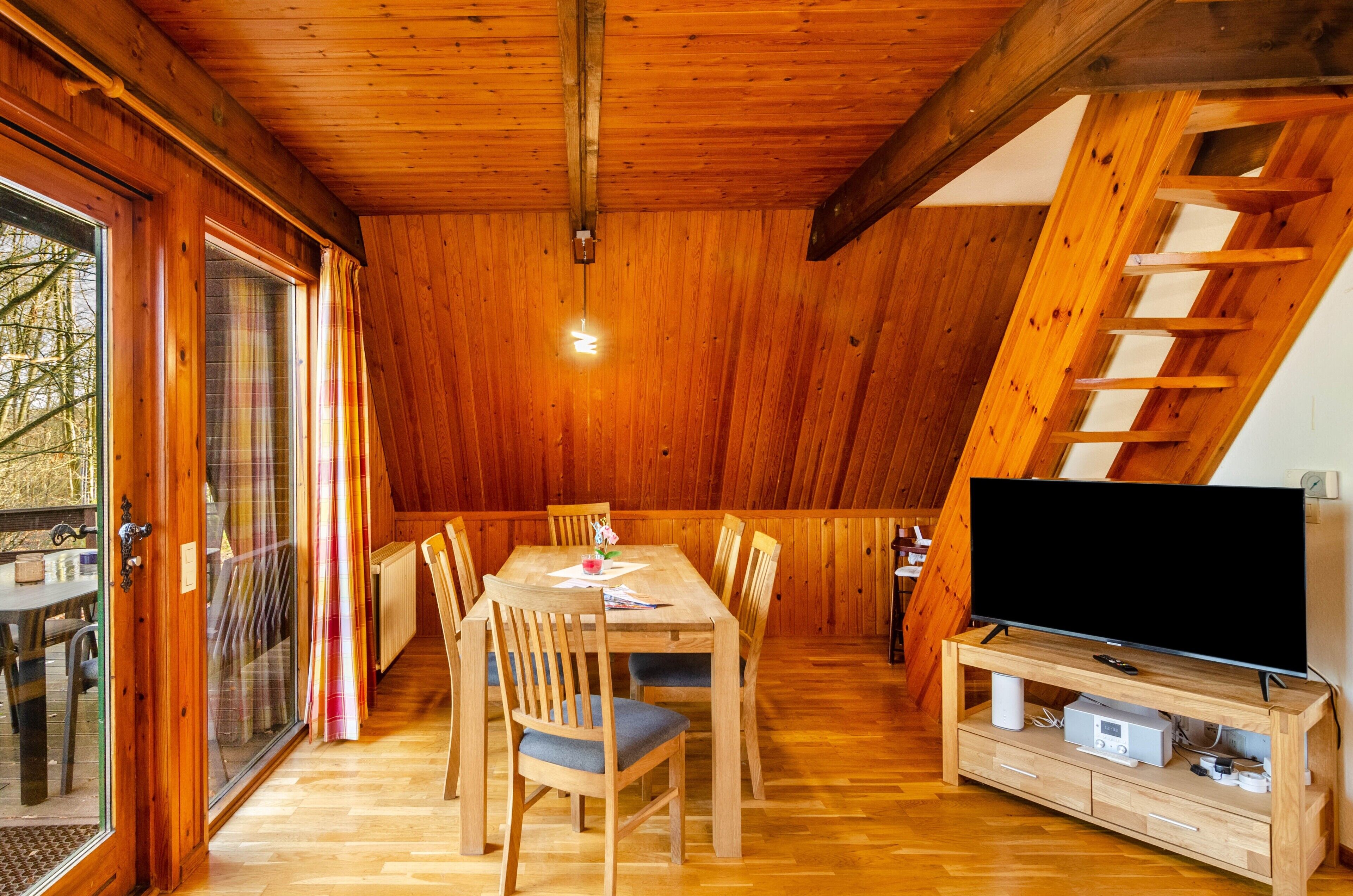 Ein Holzinterieur mit einem Esstisch, Stühlen, einem Fernseher auf einem Holzgestell und einem Fenster mit Vorhängen.