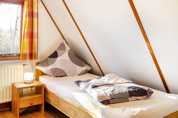 A small, cozy room with a sloped ceiling, a single bed with patterned bedding, a bedside table with a lamp, and a window with a plaid curtain.