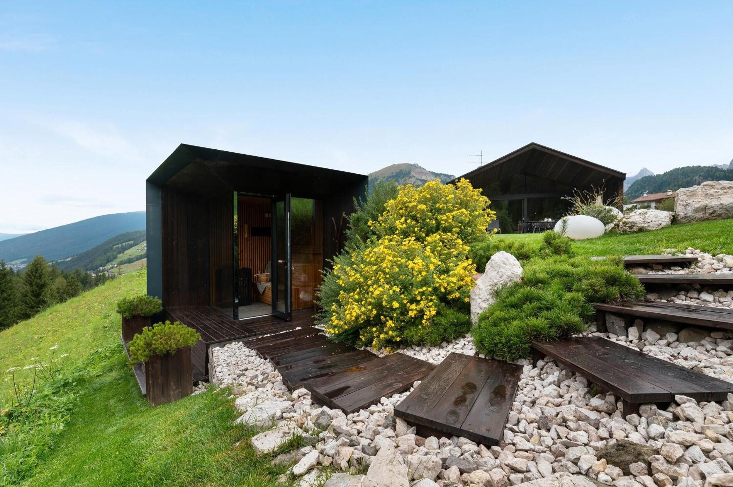Eine moderne Holzhütte mit Glastür, umgeben von einem Garten mit gelben Blumen und Steinen.