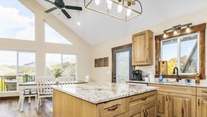 A spacious kitchen with wooden cabinets, a granite countertop, and a large island. There is a dining area with a table and chairs, and a view of the outdoors through large windows. - Lava`s Red King of The Hill (Lava Hot Springs)