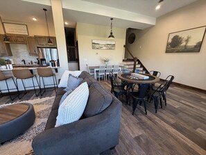 A modern living room with a gray sofa, dining table, and chairs. A kitchen is visible in the background.