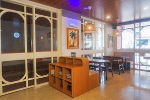 Un restaurante con mostrador de recepción de madera, mesas, sillas y grandes ventanales que permiten la entrada de luz natural.