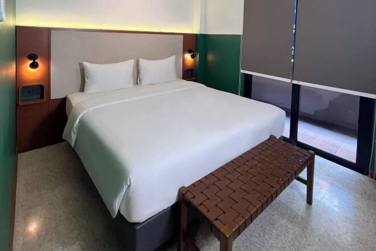 Une chambre d’hôtel avec un grand lit, deux tables de chevet avec des lampes, un banc et une vue sur l’extérieur grâce à une porte vitrée.
