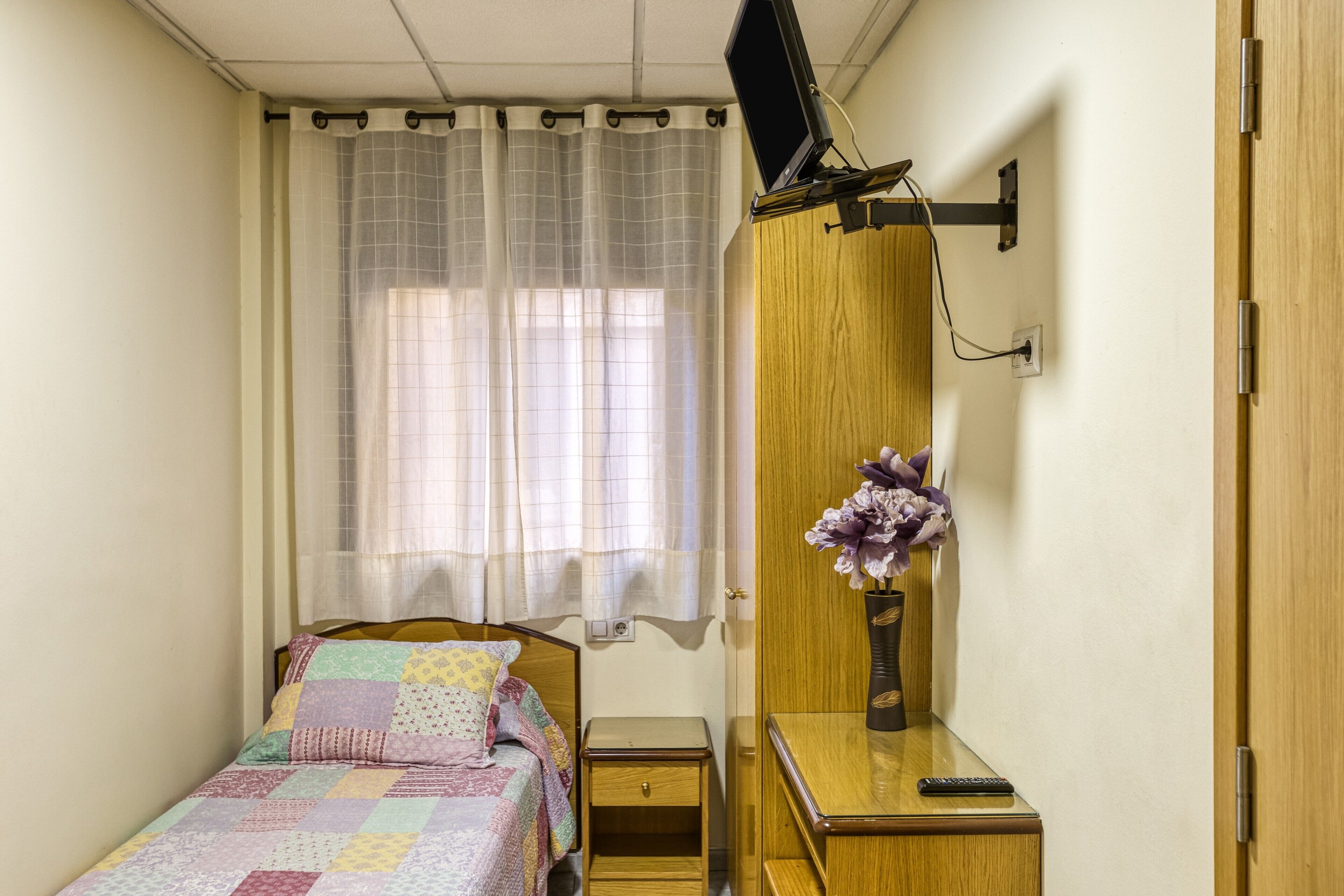 Een kleine, verzorgde slaapkamer met een bed, nachtkastje, een aan de muur bevestigde televisie en een houten garderobe.