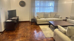A living room with a sofa, coffee table, TV, and a clock on the wall. - Spacious 4-bedroom villa with a Pool in serene Athiriver BAOB (Athi River)
