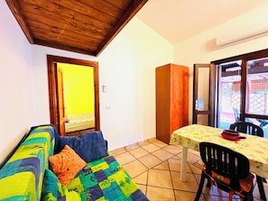 A room with a wooden ceiling, a sofa with colorful cushions, a dining table with a floral tablecloth, and a view through a window.