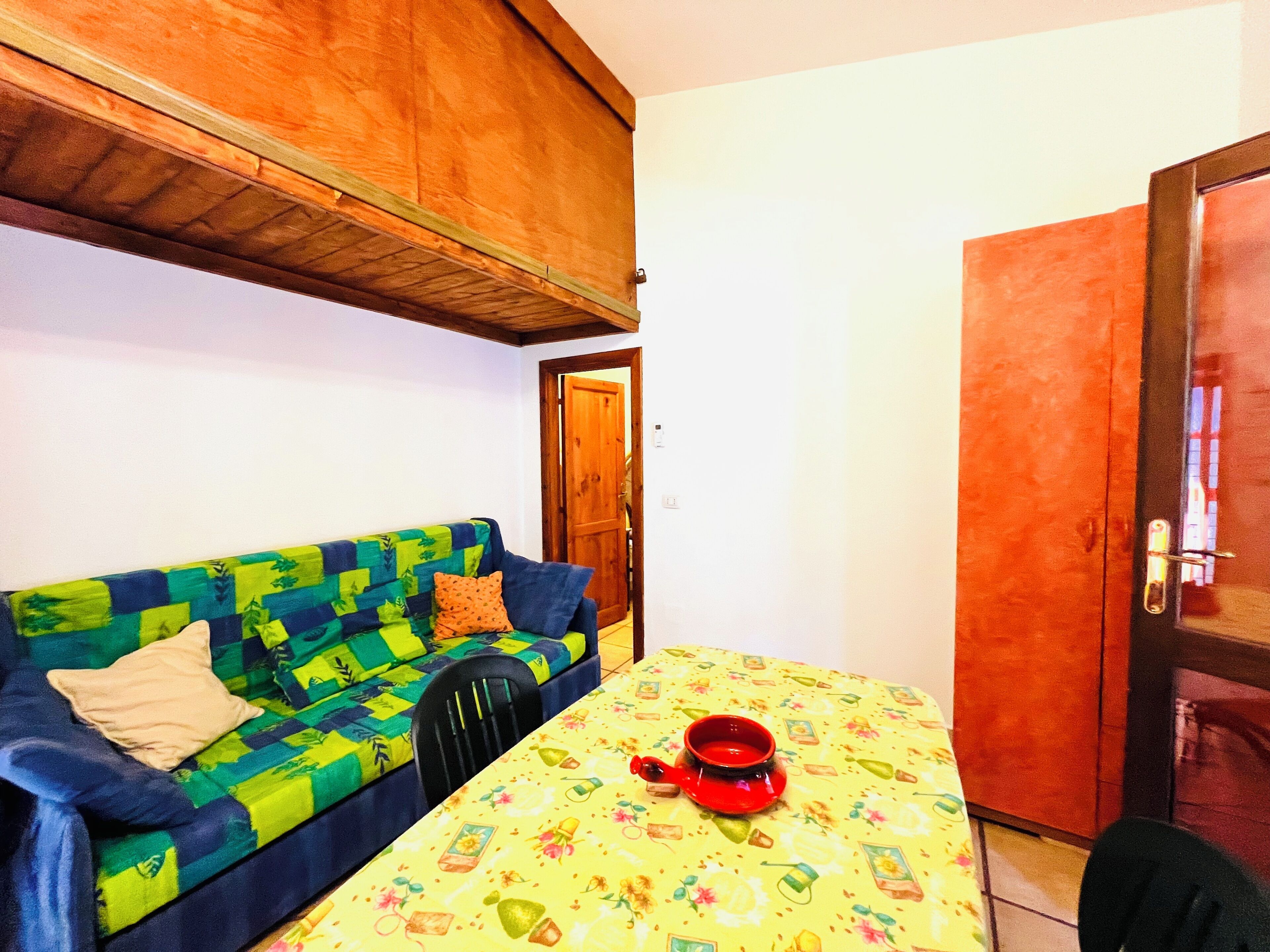 Una habitación con techo de madera, un sofá colorido, una mesa con mantel floral y una tetera roja.