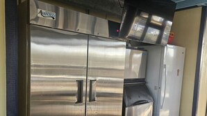 A stainless steel refrigerator and freezer unit with a vent hood, located in a covered outdoor area with a wooden floor.