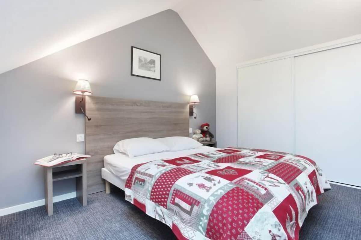 Ein Schlafzimmer mit schrägem Dach, einem hölzernen Kopfteil, zwei Nachttischlampen, einem Bett mit rotem und weißem Bettbezug, einem Nachttisch mit einem Buch und einem Teddybären.